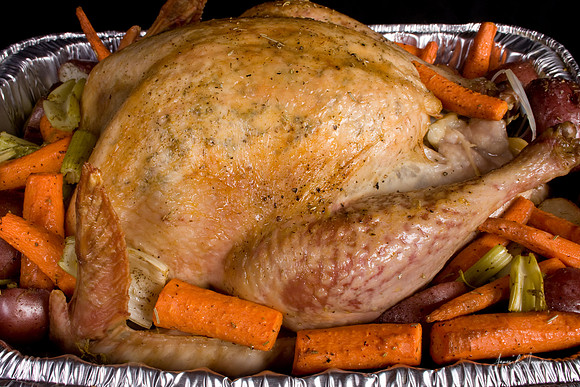 Close-up of a golden brown roasted holiday dinner whole turkey surrounded by vegetables of orange carrots, green celery and red potatoes in a silver roasting pan on a black background.