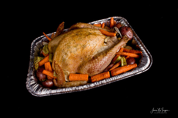 A golden brown roasted holiday dinner whole turkey surrounded by vegetables of orange carrots, green celery and red potatoes in a silver roasting pan on a black background.